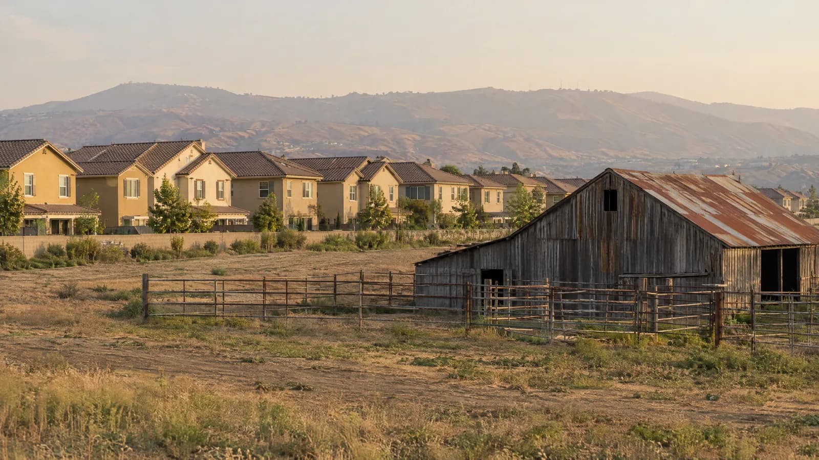 Row of new two-story stucco homes with tile roofs at the edge of a flat former dairy pasture, an old weathered wooden barn and empty corral fencing in the foreground and hazy Chino Hills on the horizon