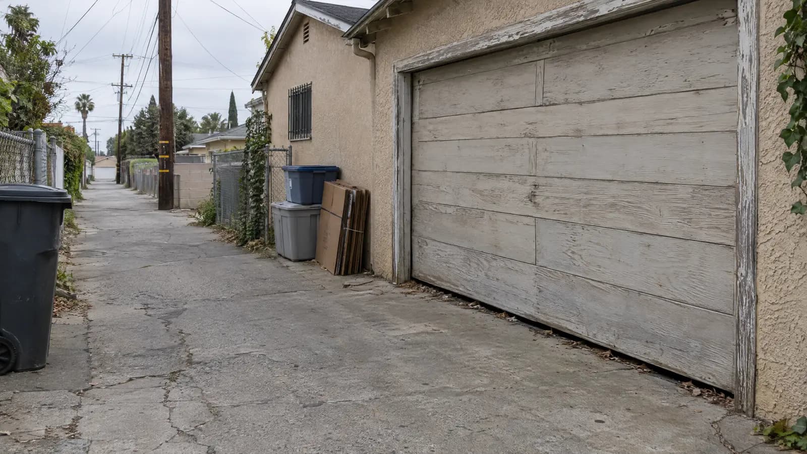 Narrow alley behind stucco houses with stacked storage bins and a visible gap under a weathered wooden garage door, a common rat entry point