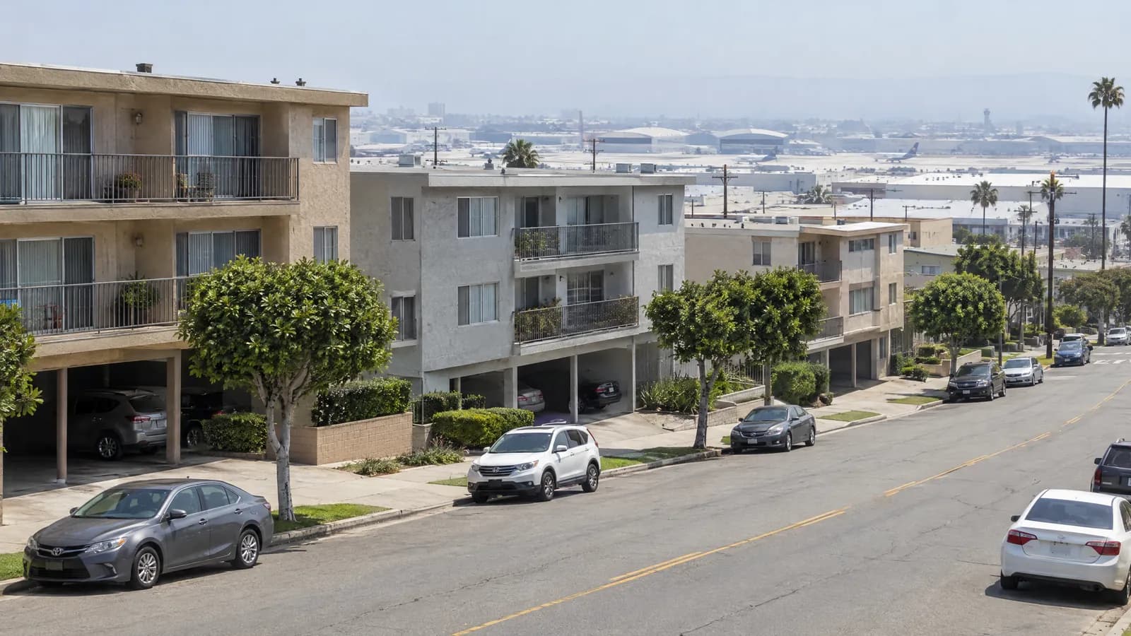 Three-story 1970s stucco apartment buildings with ground-level carports along a wide South Bay street, with the flat roofs of hangars and industrial buildings beyond under a hazy sky