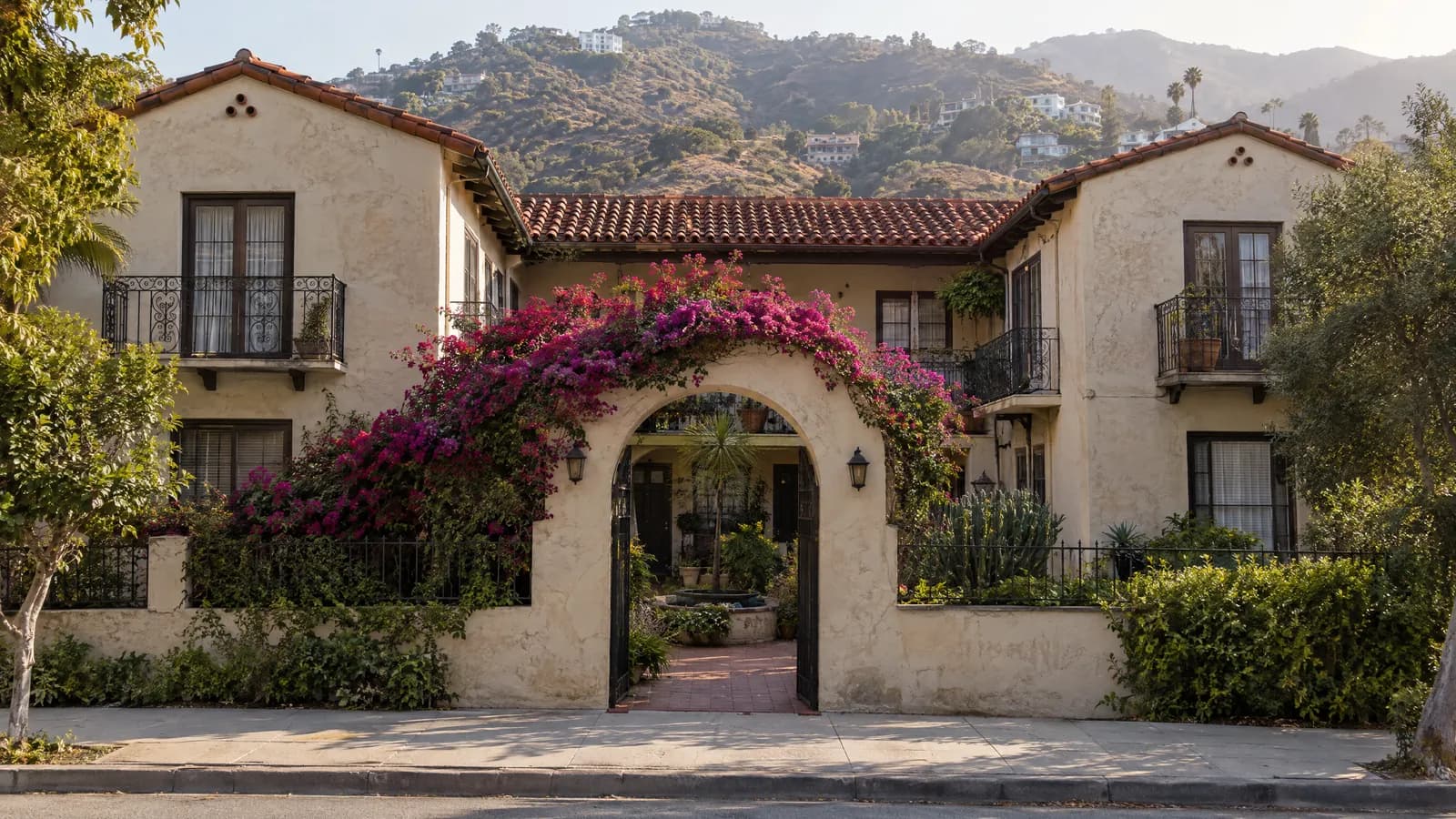 Two-story 1920s Spanish-style courtyard apartment building with cream stucco, red clay tile roof and an arched entry draped in bougainvillea, with chaparral-covered hills rising behind in hazy afternoon light
