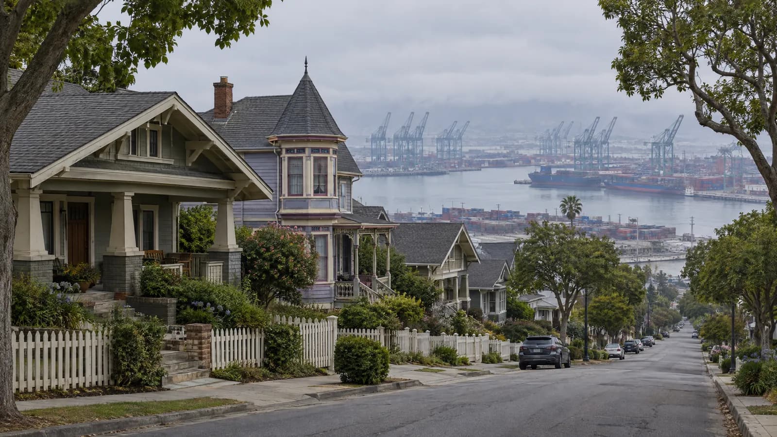 Hillside street of early-1900s wood-sided Craftsman and Victorian cottages with deep porches under a gray morning marine-layer sky, with port gantry cranes and the harbor channel visible below in the distance