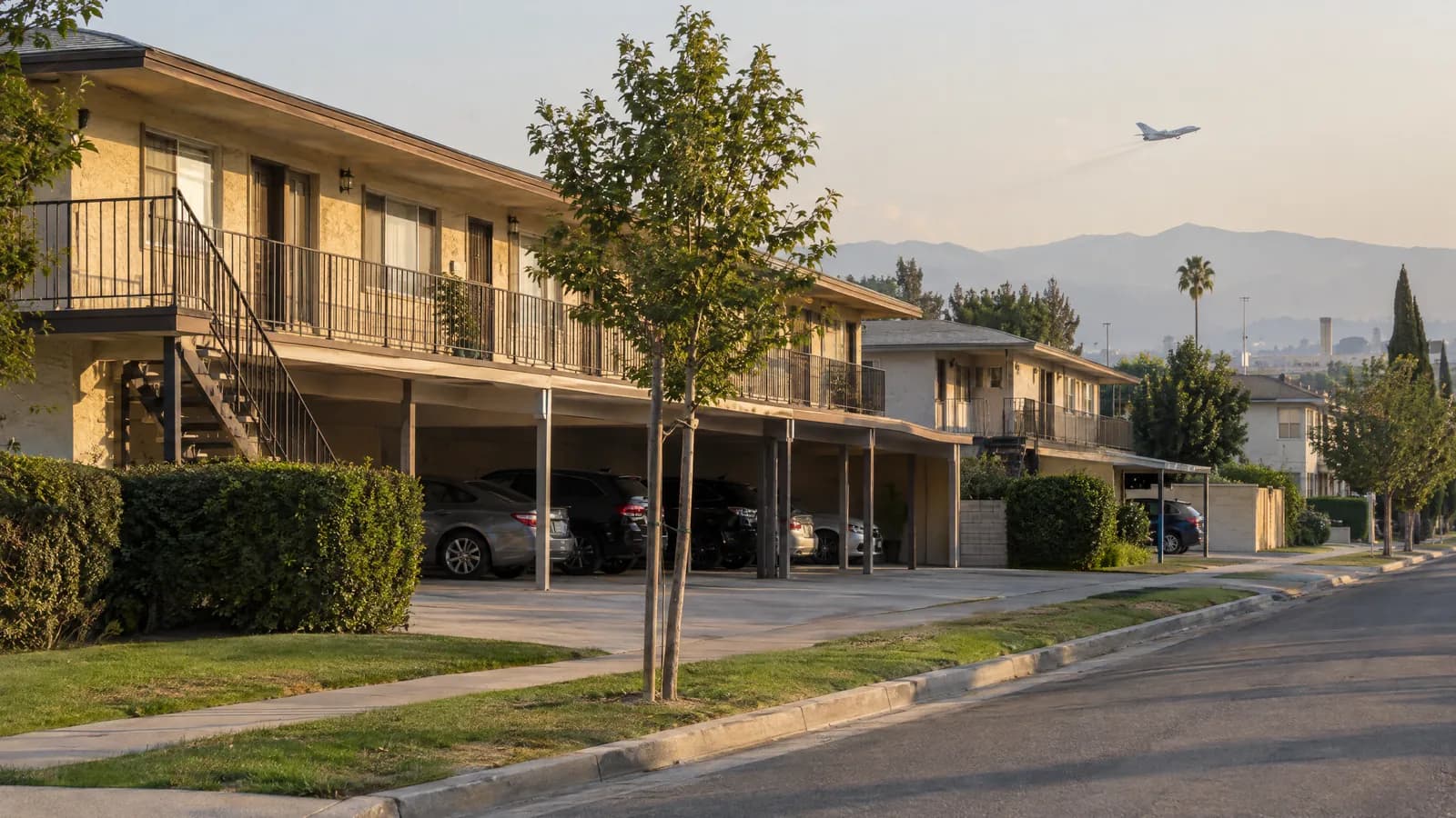 Two-story 1970s stucco garden apartment building in Van Nuys with exterior walkways, carports and a strip of lawn, as a small business jet climbs through the hazy sky beyond the rooftops